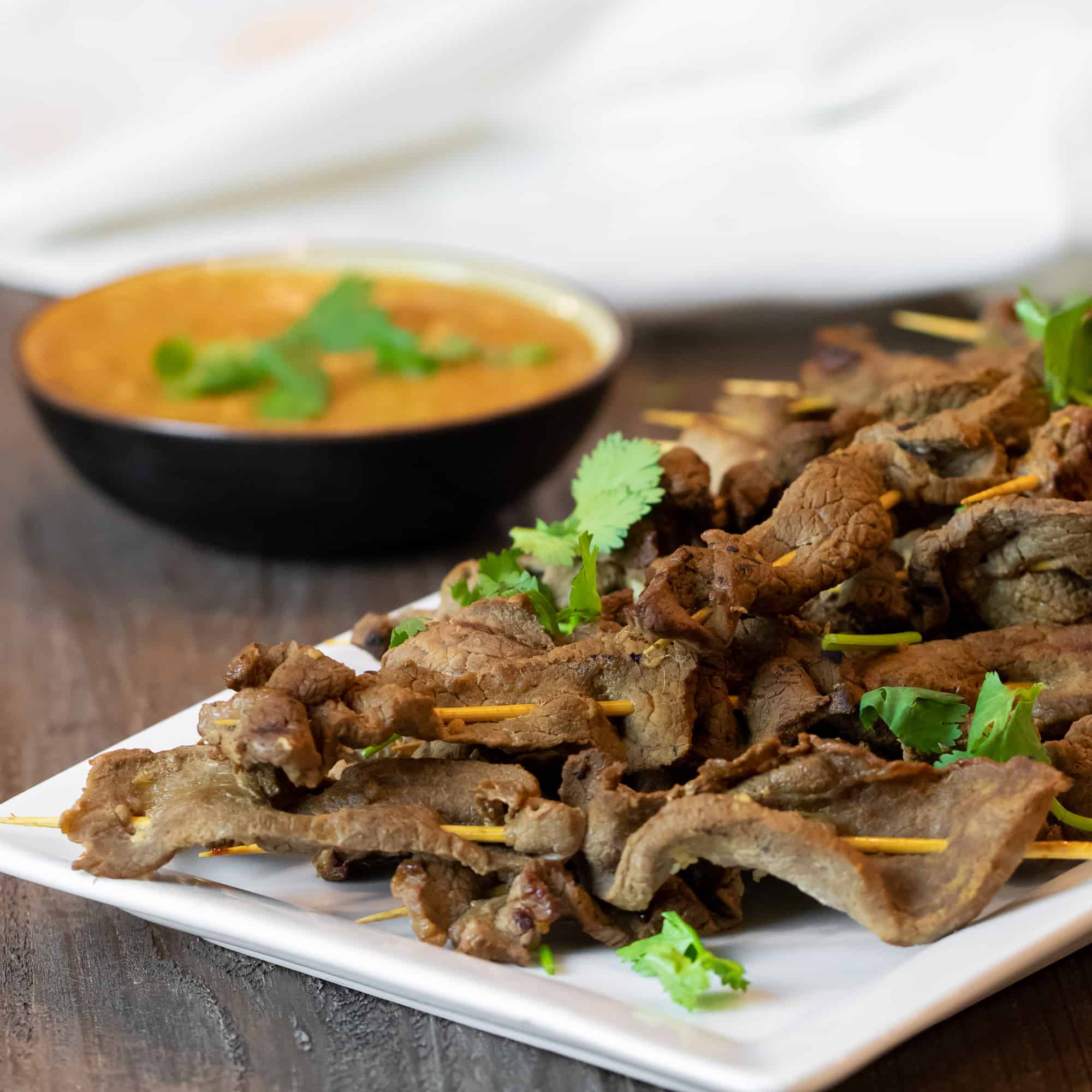 A platter of beef satay skewers next to a bowl of Thai peanut sauce.