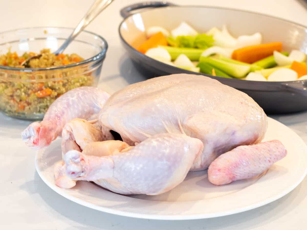 A raw chicken with a bowl of stuffing and the roasting dish in the back.