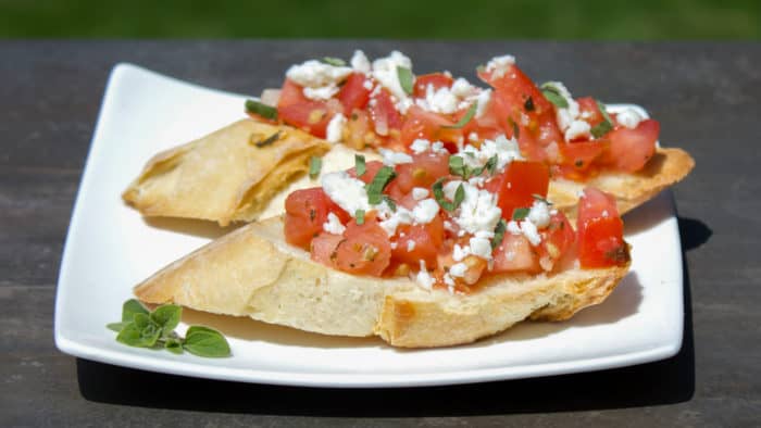 Recipe for how to make bruschetta bread with roma tomatoes, garlic, onion, lemon juice and olive oil. Topped with feta and oregano on a toasted slice of French baguette.