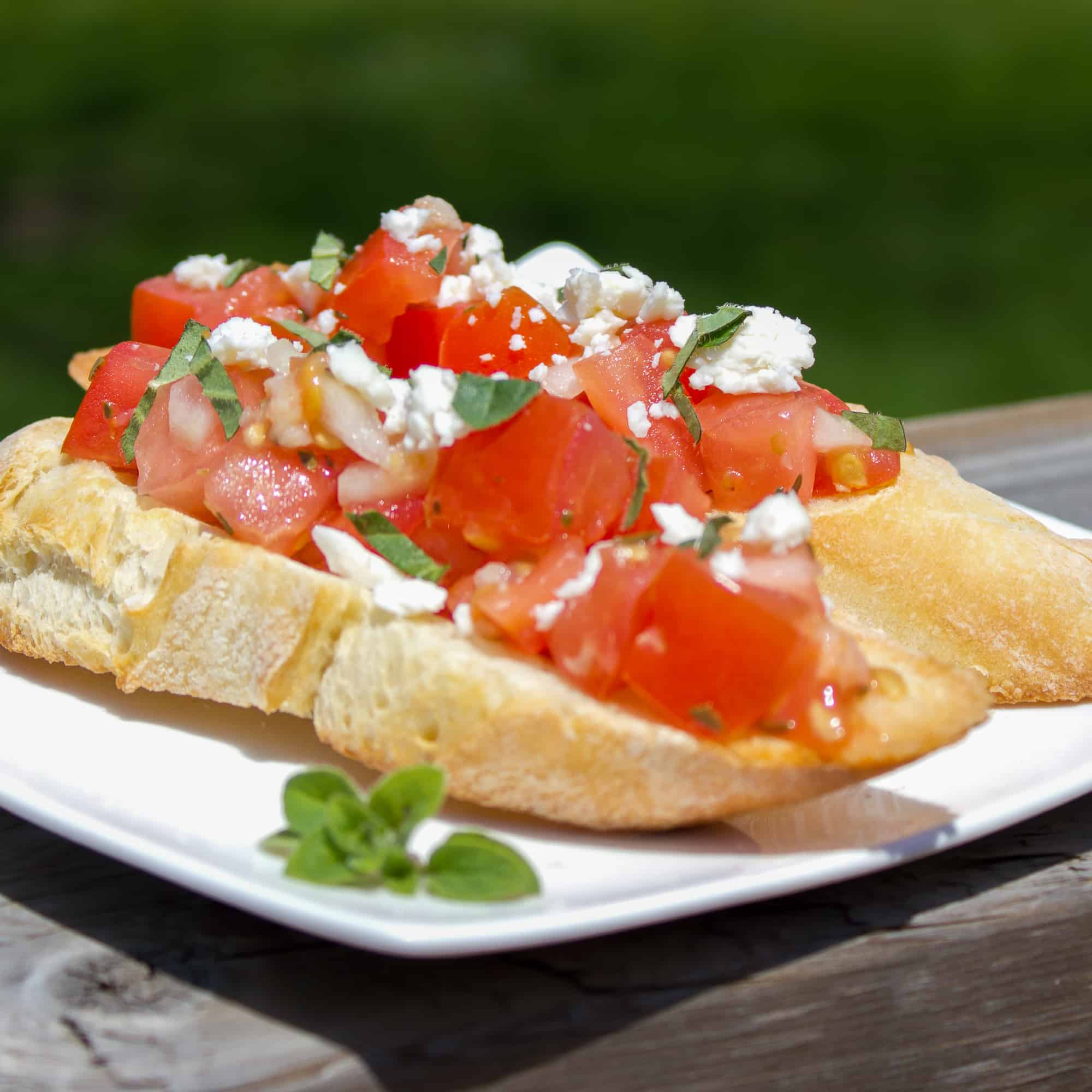 Recipe for how to make bruschetta bread with roma tomatoes, garlic, onion, lemon juice and olive oil. Topped with feta and oregano on a toasted slice of French baguette.