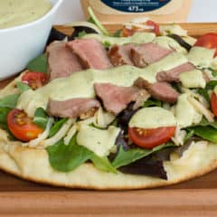 A grilled naan pizza with steak, grape tomatoes, mixed greens, pepper jack cheese and an avocado ranch dressing with cilantro, lime and hidden valley ranch spicy.
