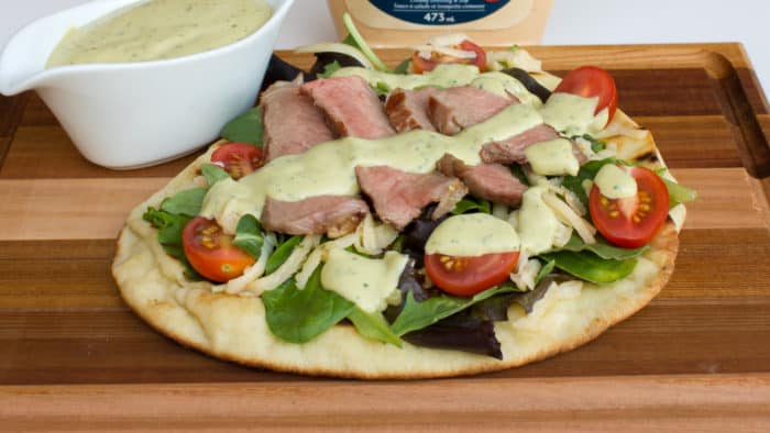 A grilled naan pizza with steak, grape tomatoes, mixed greens, pepper jack cheese and an avocado ranch dressing with cilantro, lime and hidden valley ranch spicy.