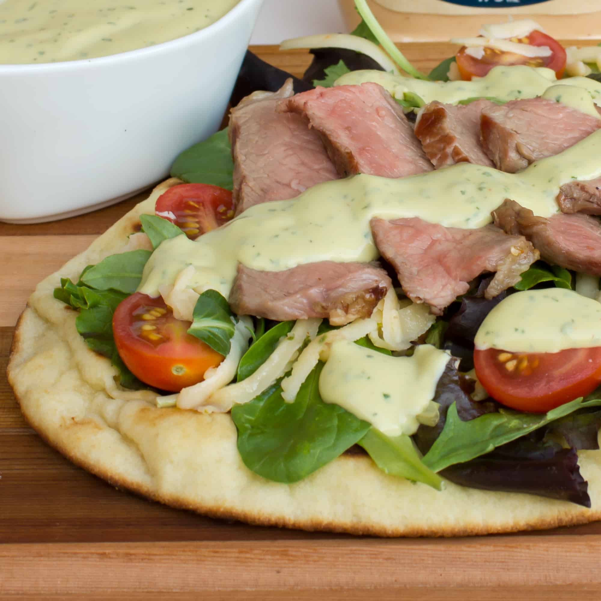 A grilled naan pizza with steak, grape tomatoes, mixed greens, pepper jack cheese and an avocado ranch dressing with cilantro, lime and hidden valley ranch spicy.