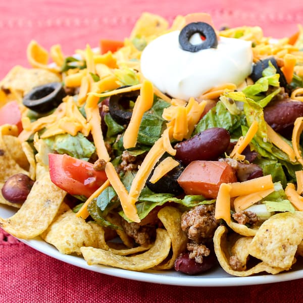 Taco salad recipe with romaine lettuce, ground beef, tomatoes, kidney beans, black olives, cheddar cheese, sour cream and Frito corn chips or nacho chips.