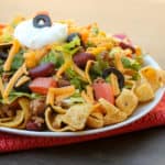 Taco salad recipe with romaine lettuce, ground beef, tomatoes, kidney beans, black olives, cheddar cheese, sour cream and Frito corn chips or nacho chips.