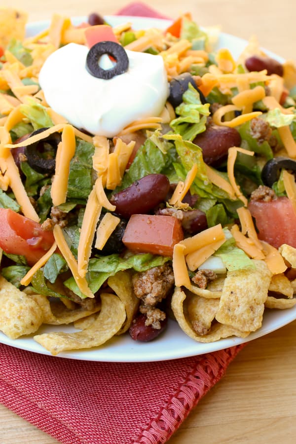 Taco salad recipe with romaine lettuce, ground beef, tomatoes, kidney beans, black olives, cheddar cheese, sour cream and Frito corn chips or nacho chips.