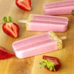 Three popsicles lying on a cutting board surrounded by strawberries.