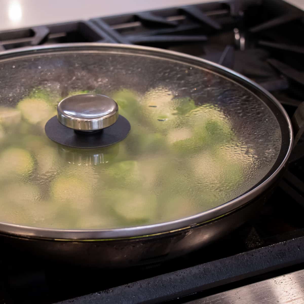 A frying pan covered with a glass lid that shows Brussels sprouts in the pan through the glass.