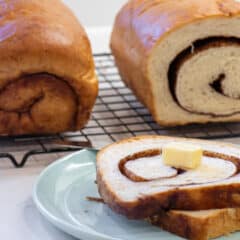 Sliced bread on a plate in front of two loaves on a wire rack.