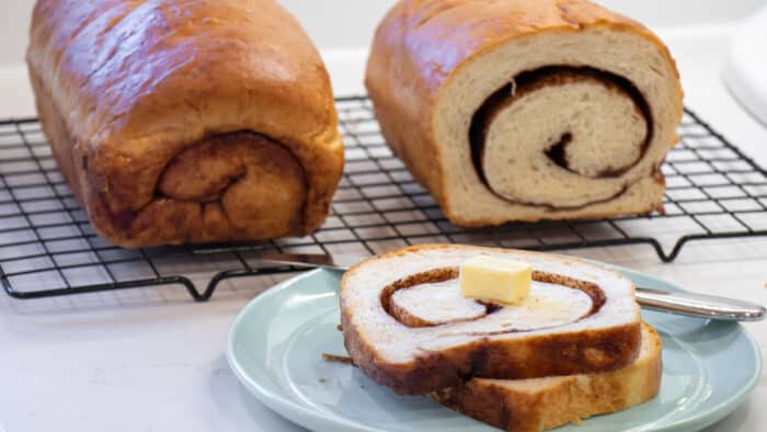 Sliced bread on a plate in front of two loaves on a wire rack.