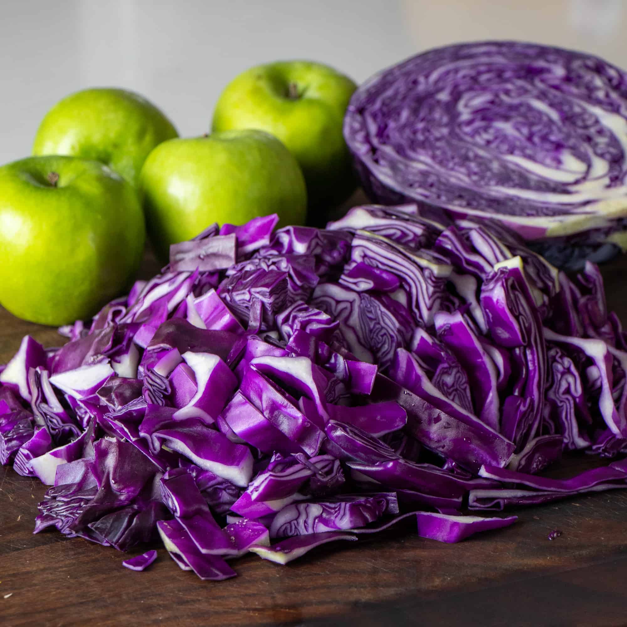 A cutting board with chopped up fresh red cabbage with granny smith apple behind it.