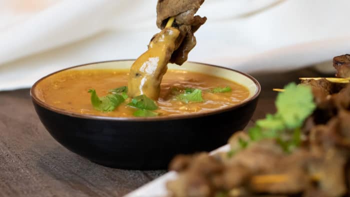 Dipping a beef satay skewer in a bowl of Thai peanut sauce