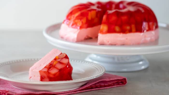 A Jello mold ring with fruit salad on a cake stand with a slice on a plate.