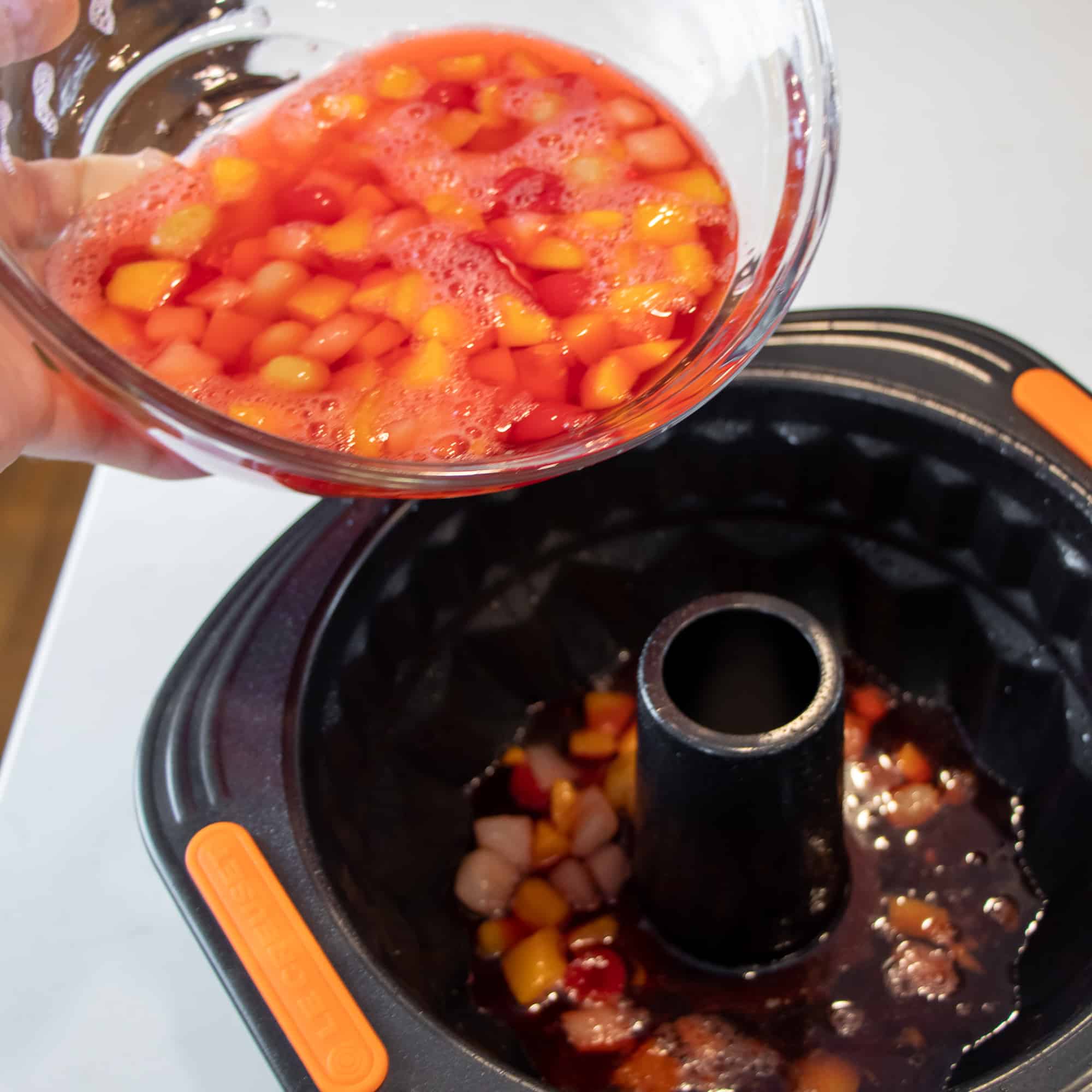 A bowl with half the jello and mixed fruit being poured into a bundt pan.