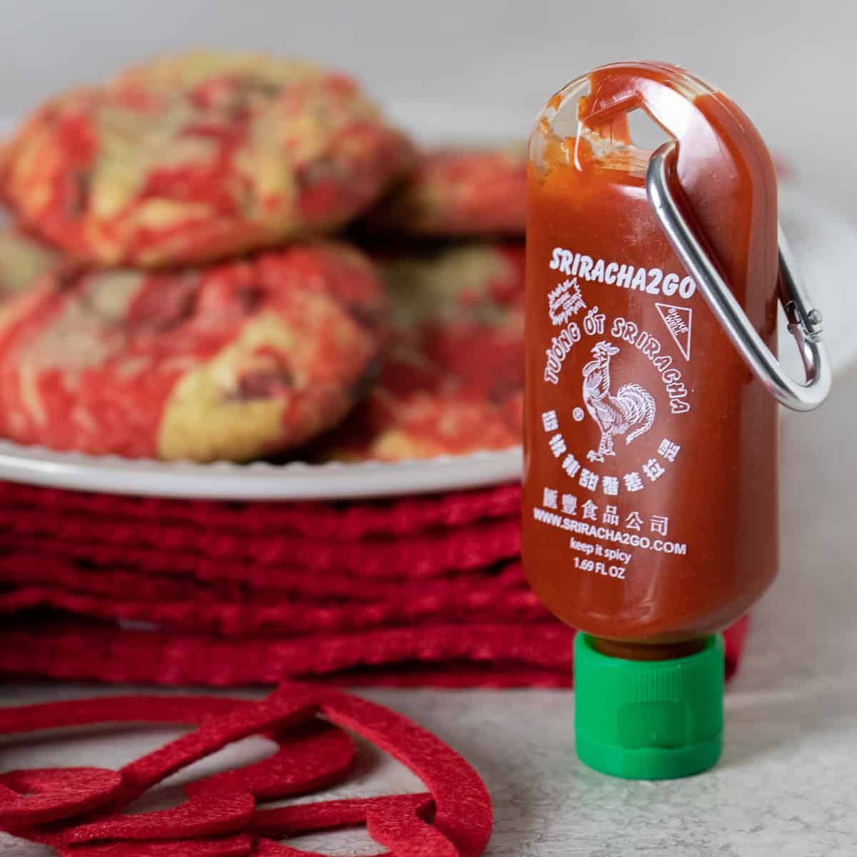 A close up picture of a bottle of Sriracha in front of a plate of chocolate chip cookies.