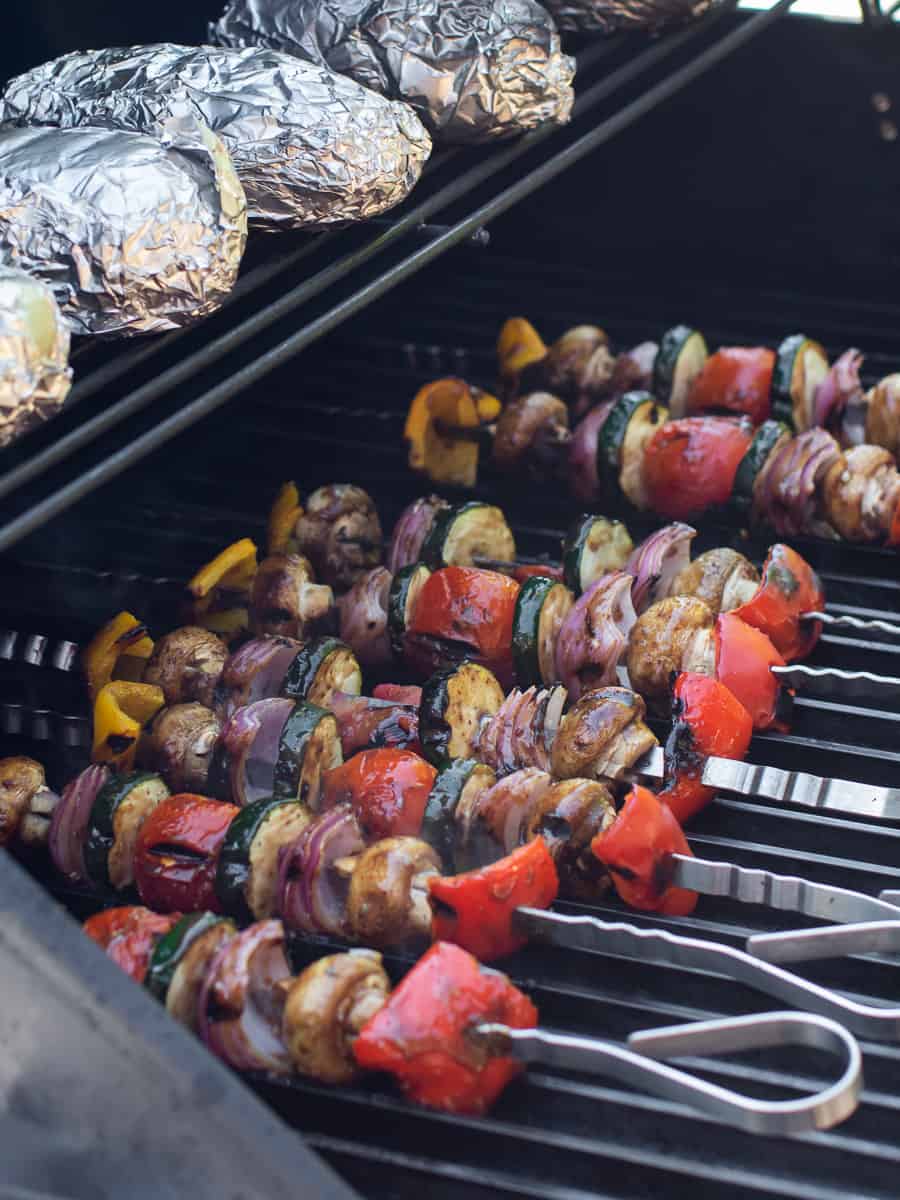 A tall image of a hot grill with potatoes and veggie skewers.