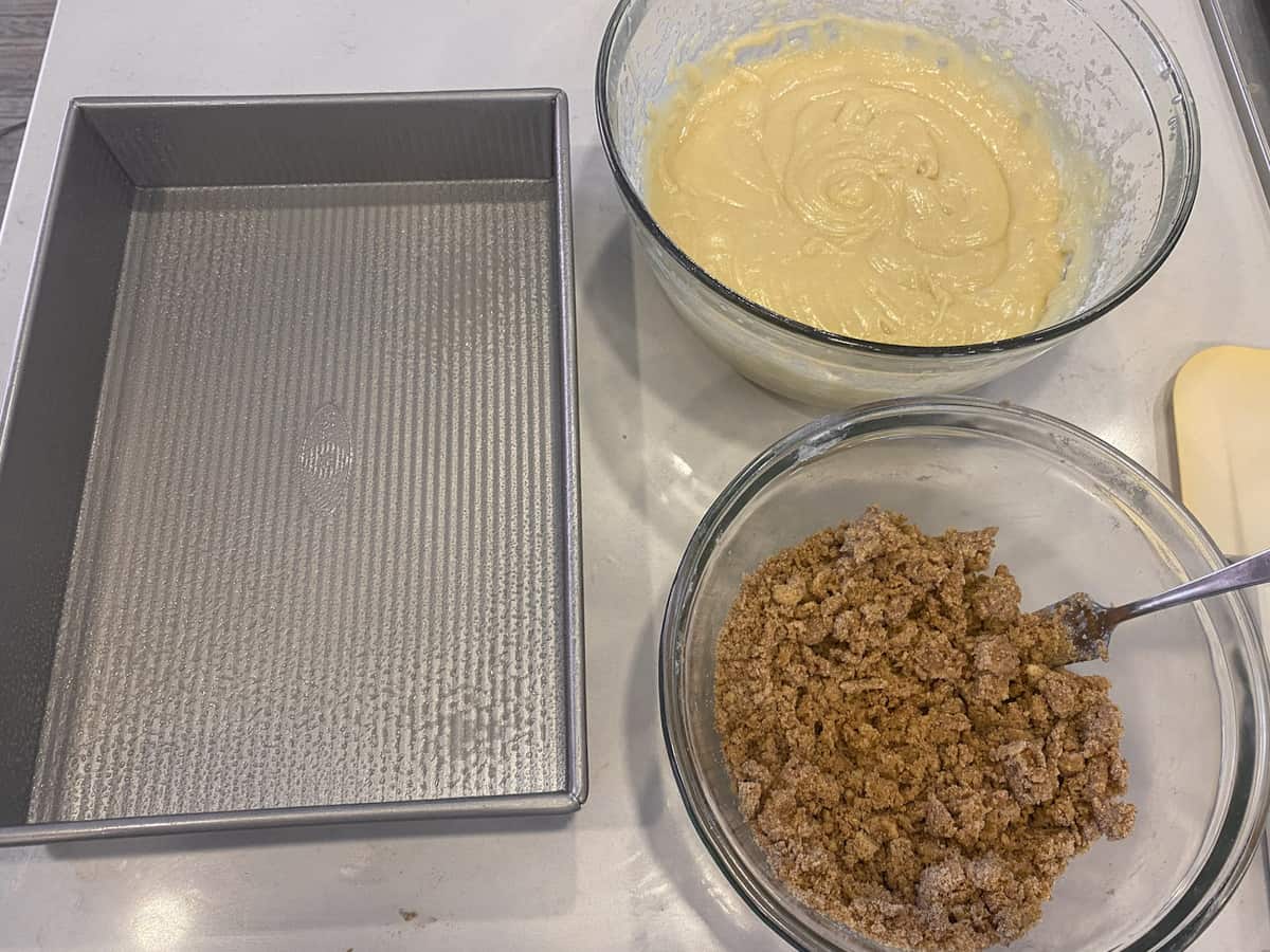 A greased rectangular baking pan with bowls of mixed batter and crumble.