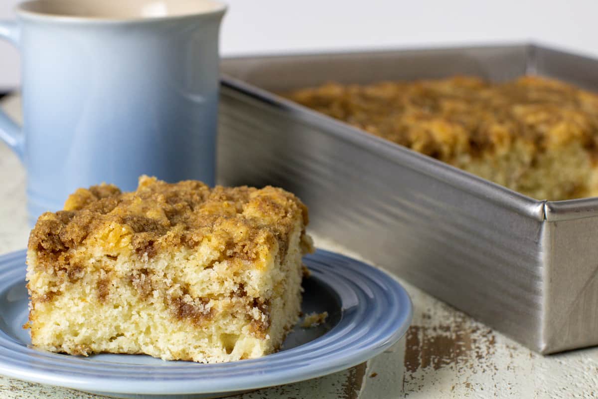 A slice of cake on a plate in front of a warm cup of coffee.