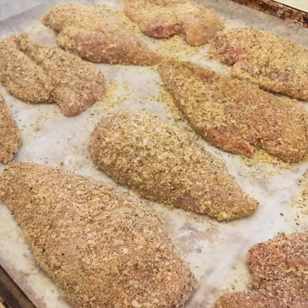 Raw chicken breast coated with bread crumbs on a baking sheet.
