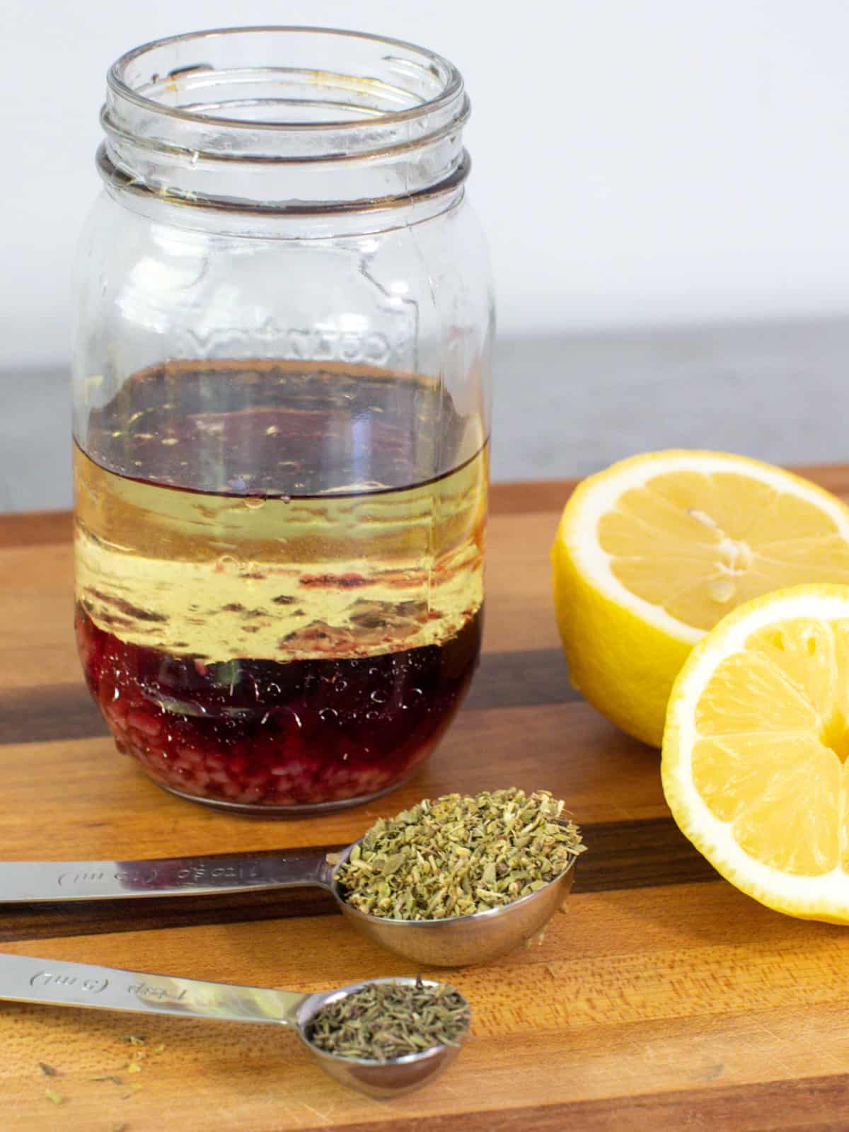 Oil and vinegar in a mason jar next to a sliced lemon and spoonfuls of oregano and thyme.