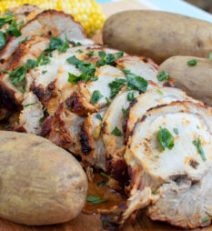 A serving board with sliced pork roast, baked potatoes and corn on the cob.