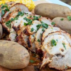 A serving board with sliced pork roast, baked potatoes and corn on the cob.