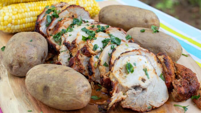 A serving board with sliced pork roast, baked potatoes and corn on the cob.