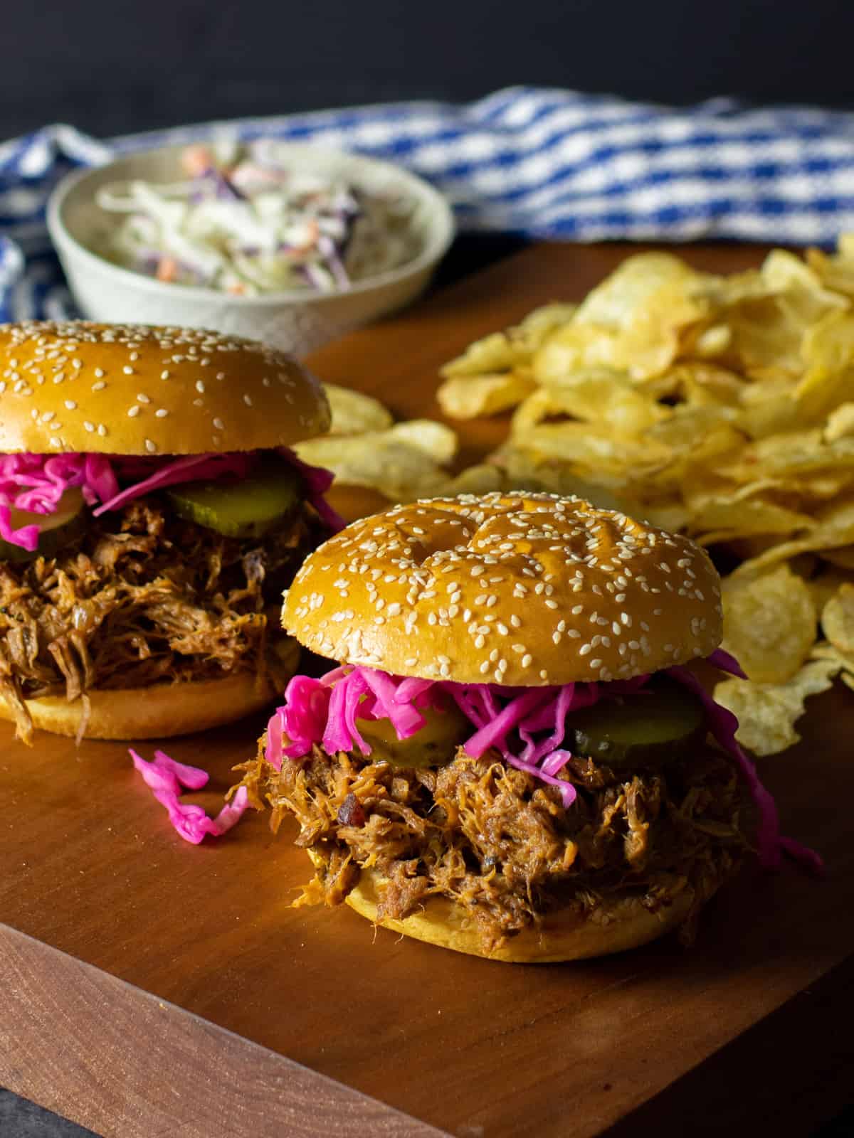 Pulled pork sandwiches on a cutting board with potato chips and cole slaw.