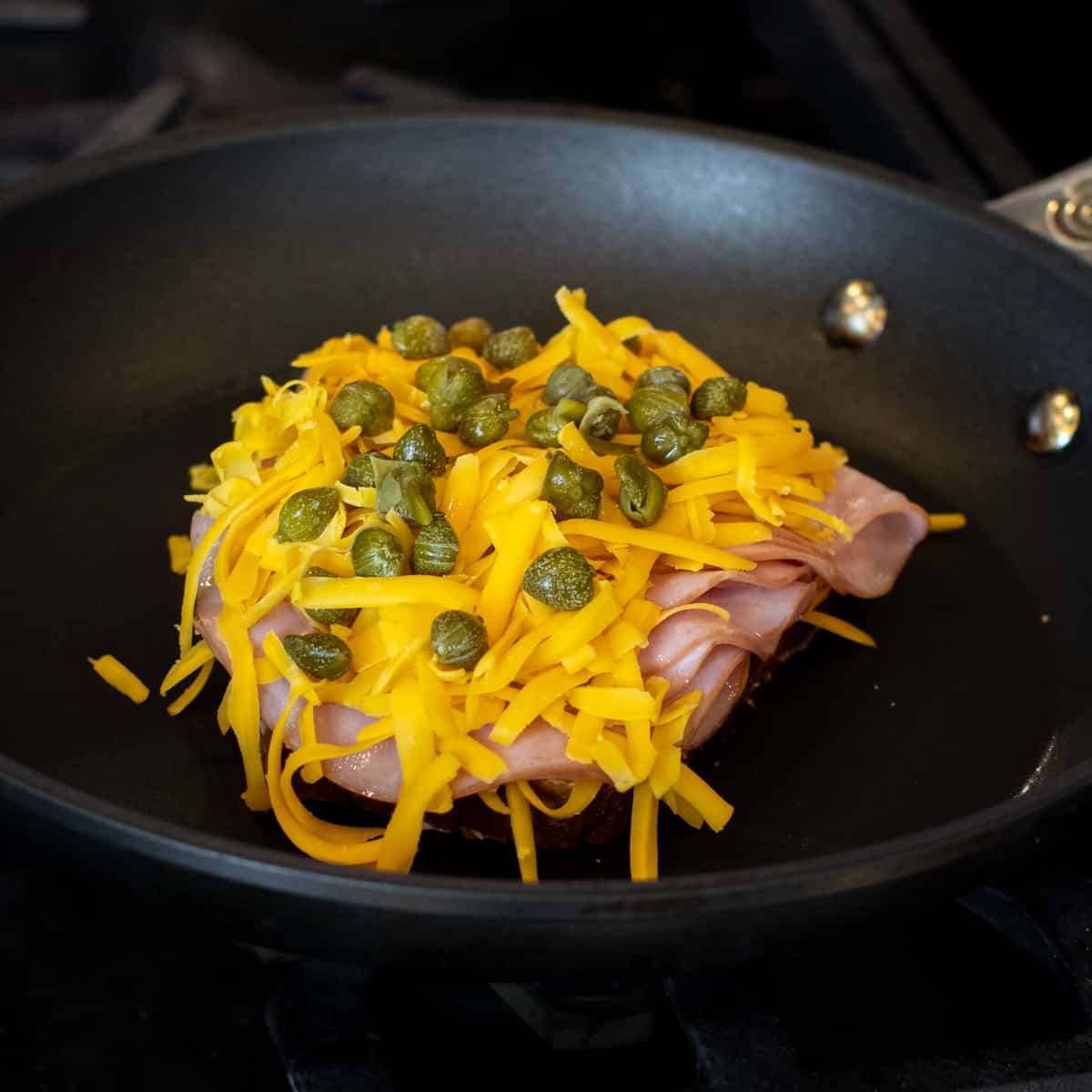 A slice of bread with ham, cheese and capers on a frying pan.