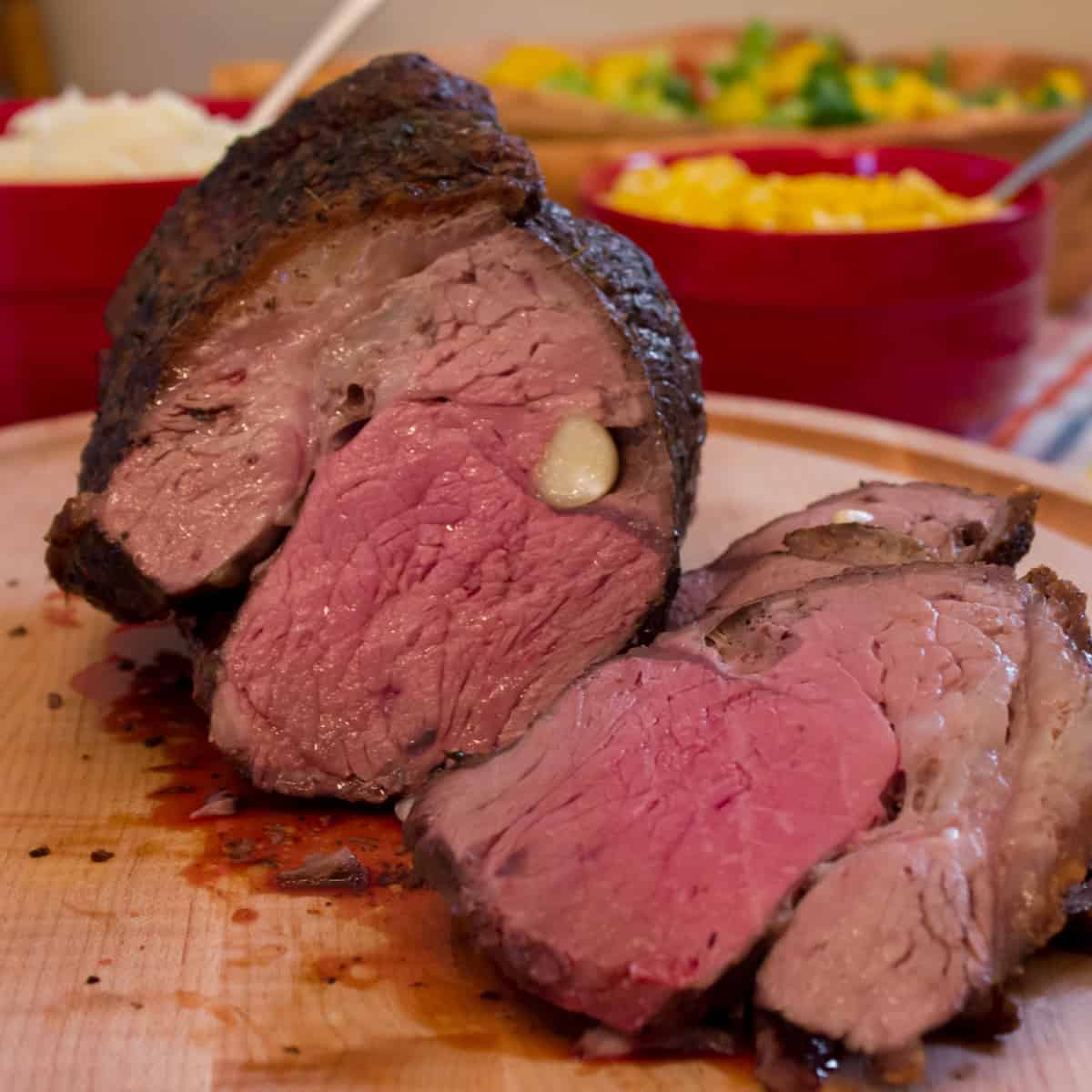 A sliced beef roast with mashed potatoes and corn in the background.