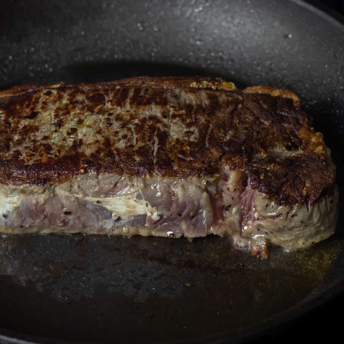 Steak searing in a frying pan with brown seared side showing on top.