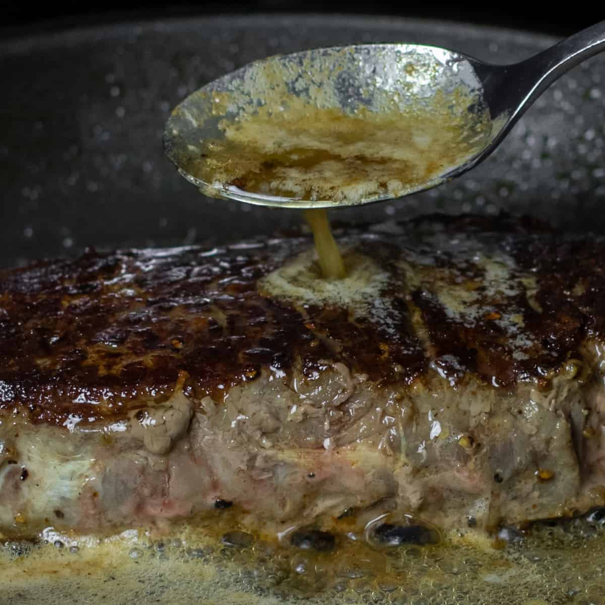 Melted butter being spooned over a steak cooking in a skillet.