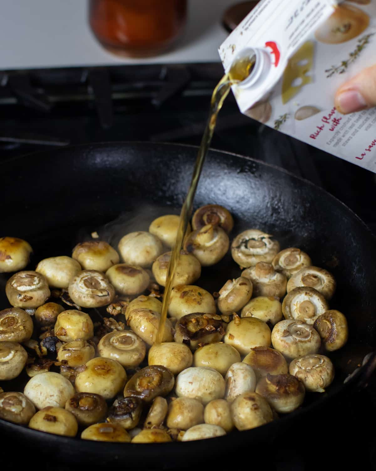 Beef broth being poured into a skillet with cooked mushrooms.