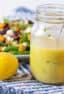 A jar of homemade lemon salad dressing in front of a plate of fresh salad.