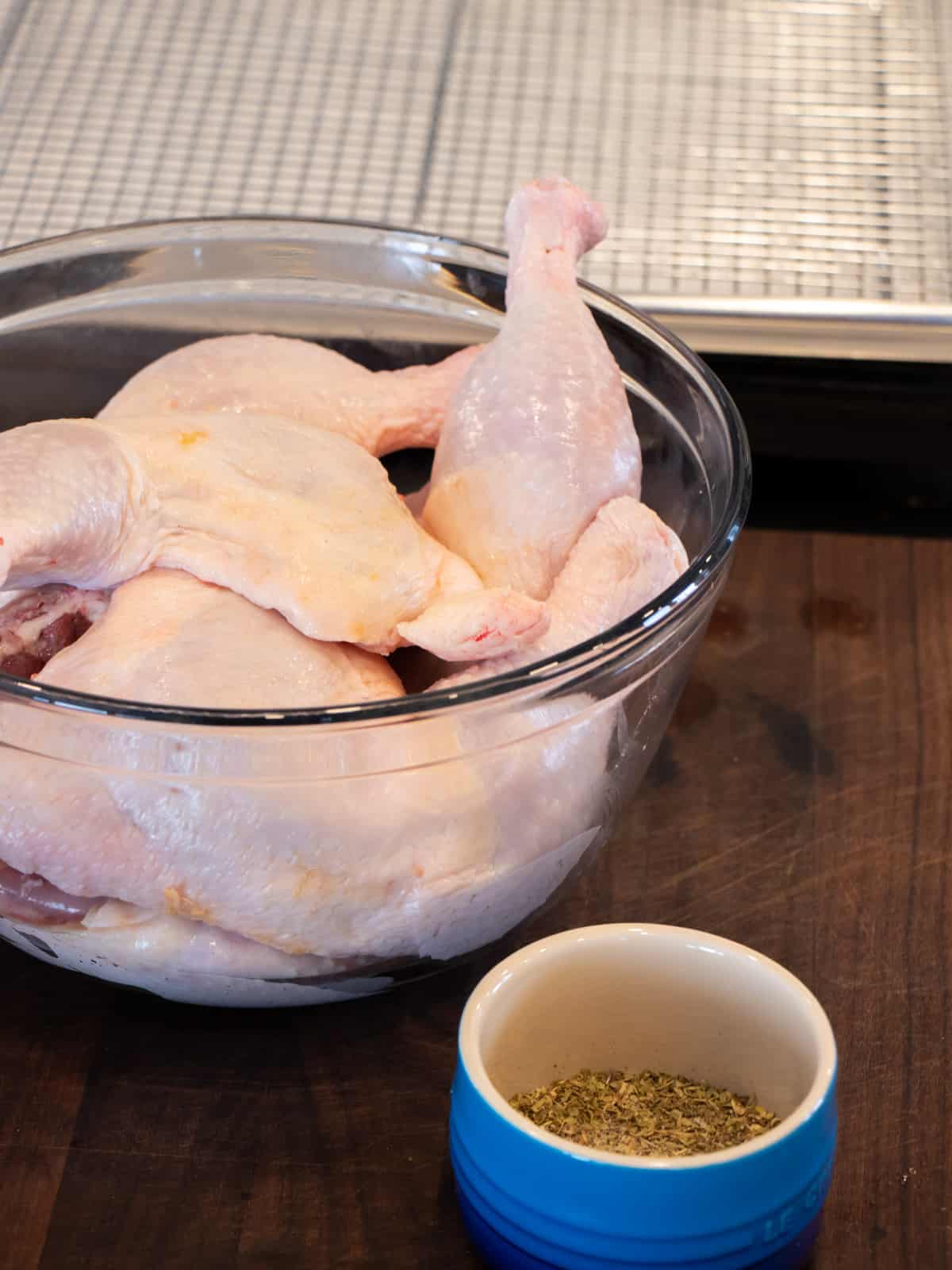 Raw chicken leg quarters in a glass bowl next to a smaller bowl filled with spices.