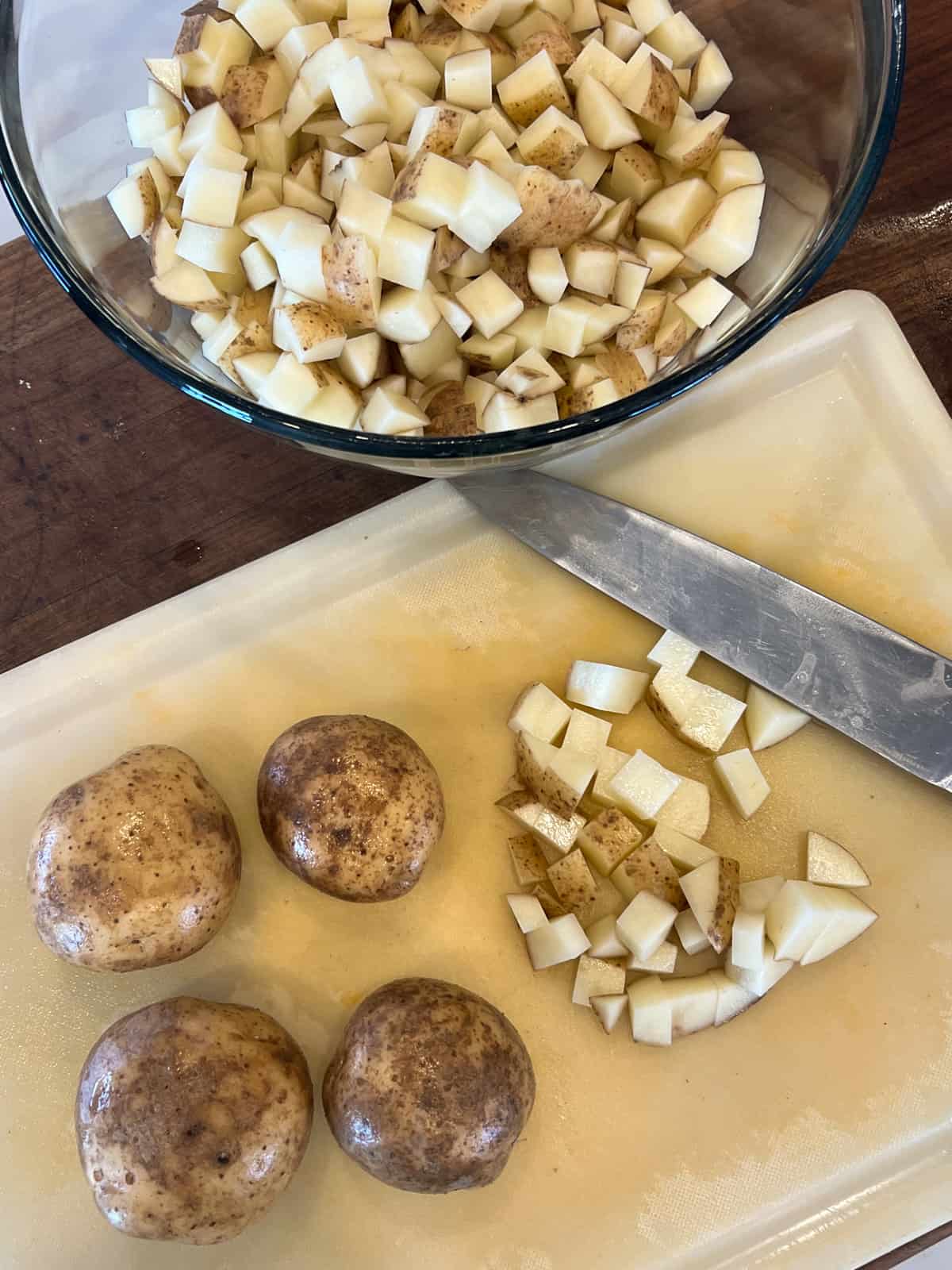 Diced potatoes in a glass bowl next to a cutting board with a knife and potatoes partially chopped.
