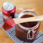 A jar of homemade BBQ sauce with a brush on top and can of pop beside it.