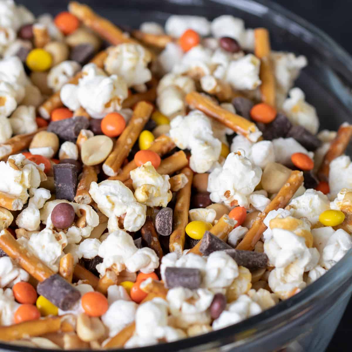 A glass mixing bowl with popcorn, pretzel sticks, chocolate and candy.