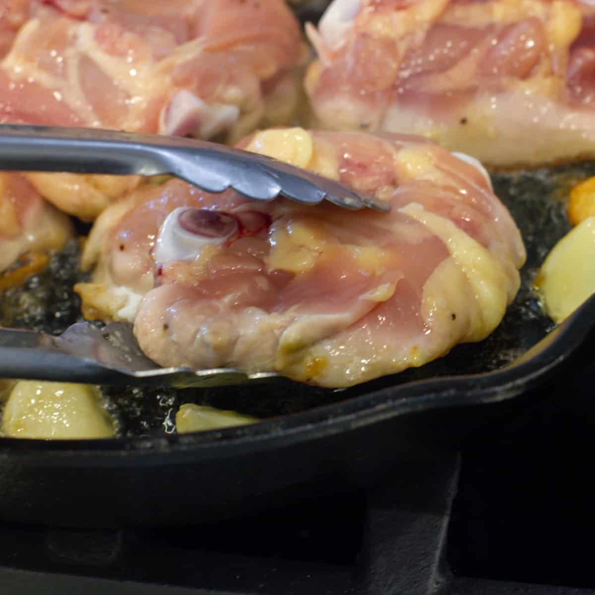 A pair of tongs flipping over a piece of chicken in a frying pan.