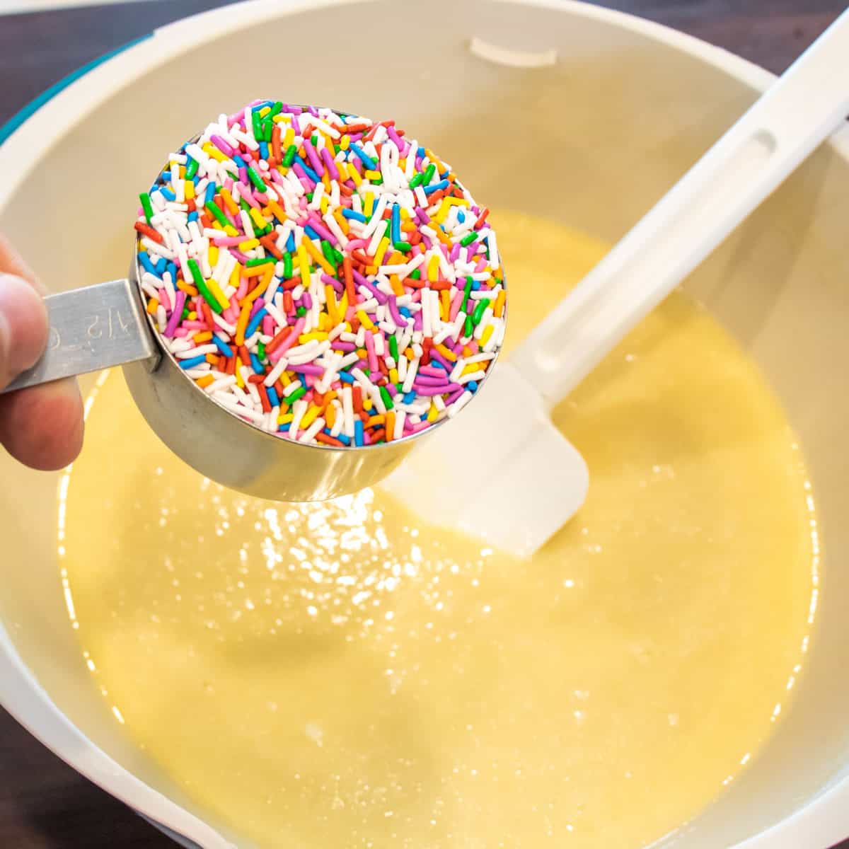 A measuring cup of sprinkles about to be poured into a bowl of cake batter.