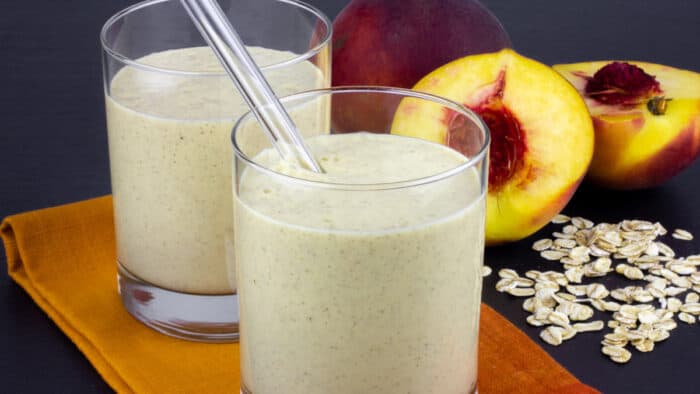 Two glasses of smoothie with halved peaches behind.