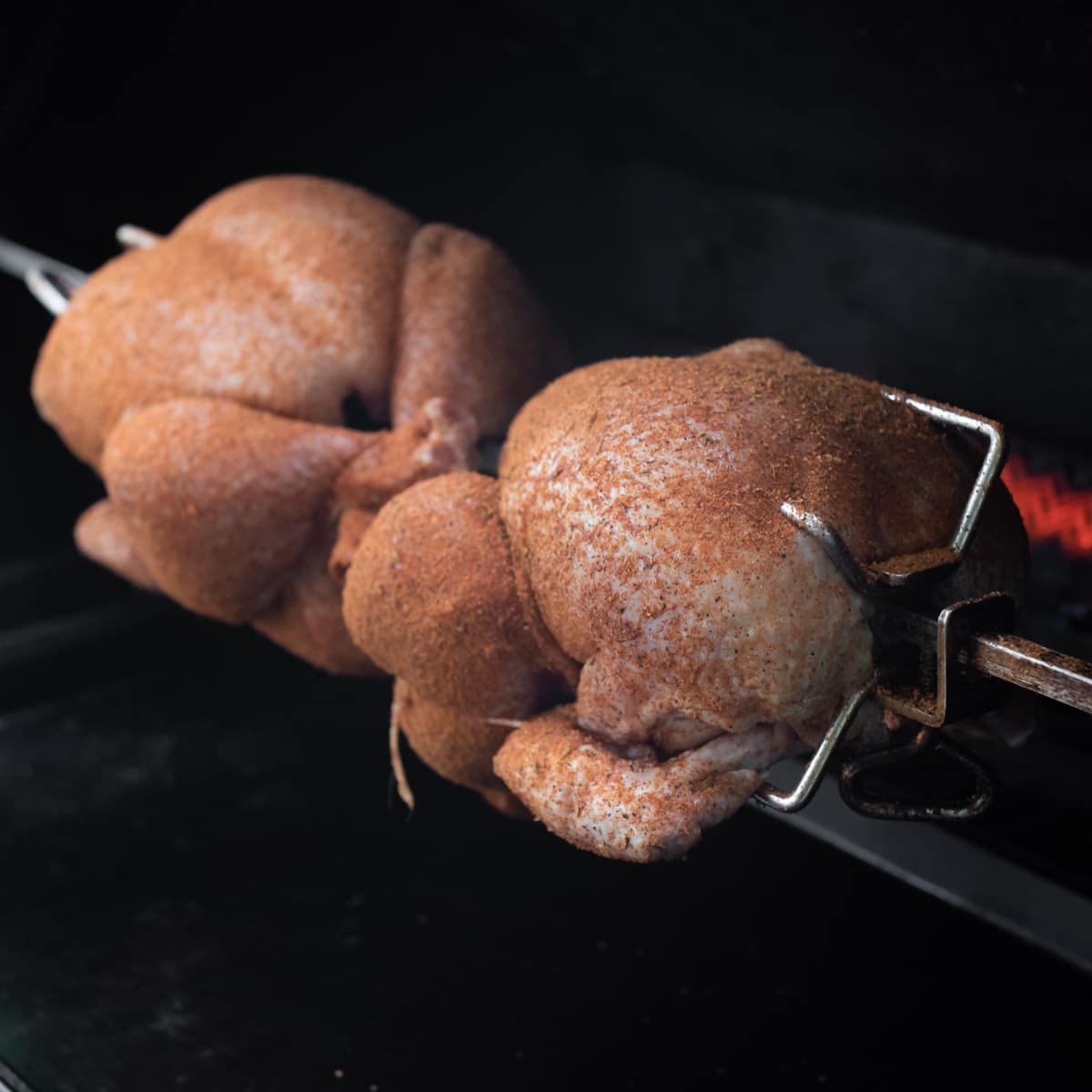 Raw seasoned chickens on a rotisserie spit with the flame in the background.