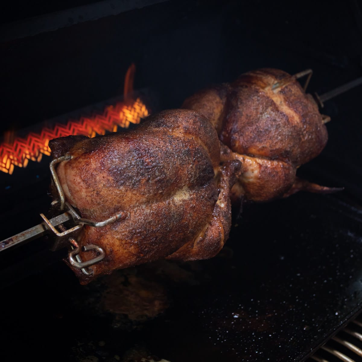 Cooked chickens turning on a spit on the grill with the flame behind them.