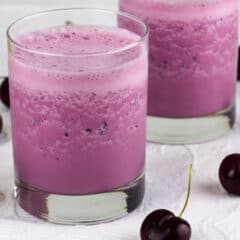 Black cherries on a white placemat with two glasses filled with a drink.
