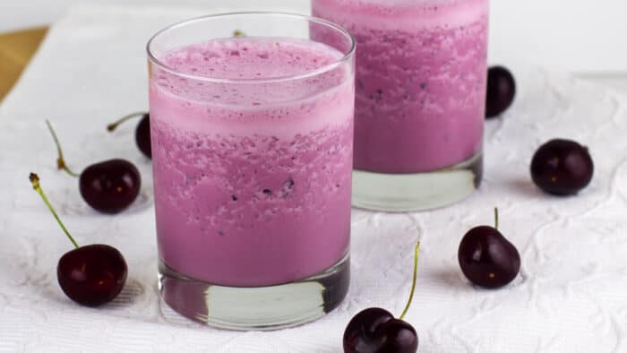 Black cherries on a white placemat with two glasses filled with a drink.