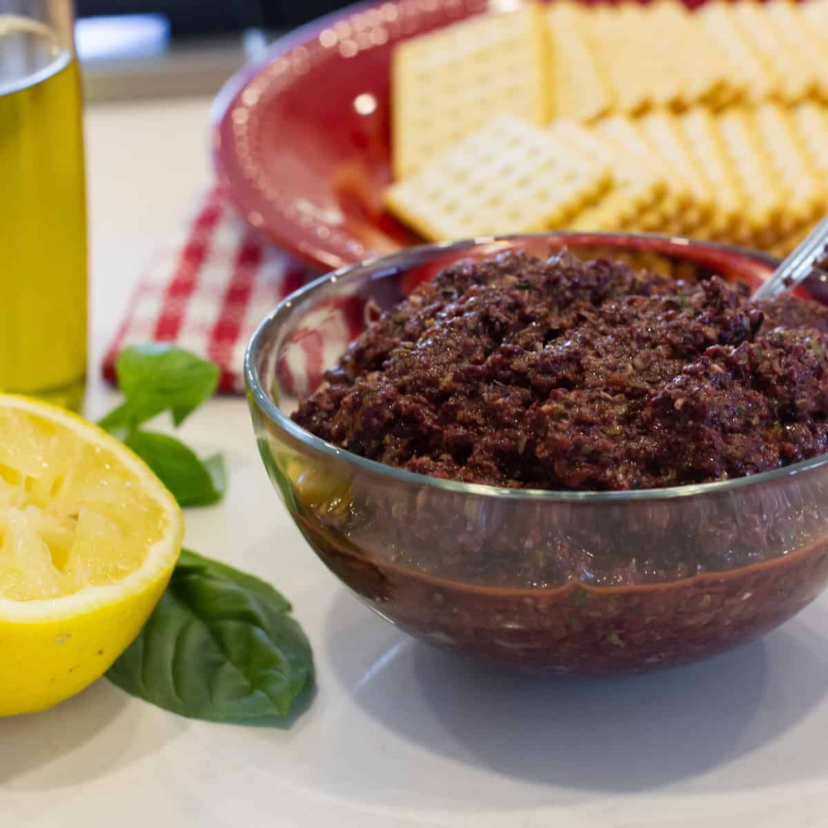 A bowl of tapenade with a plate of crackers behind.