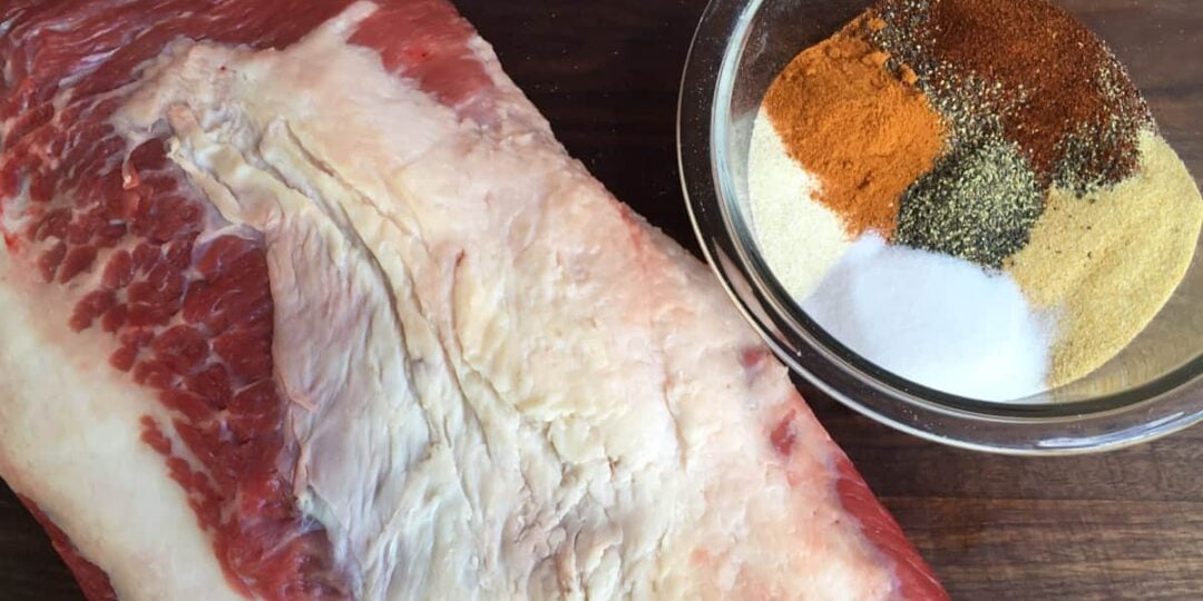 A bowl of spices next to a large beef roast.