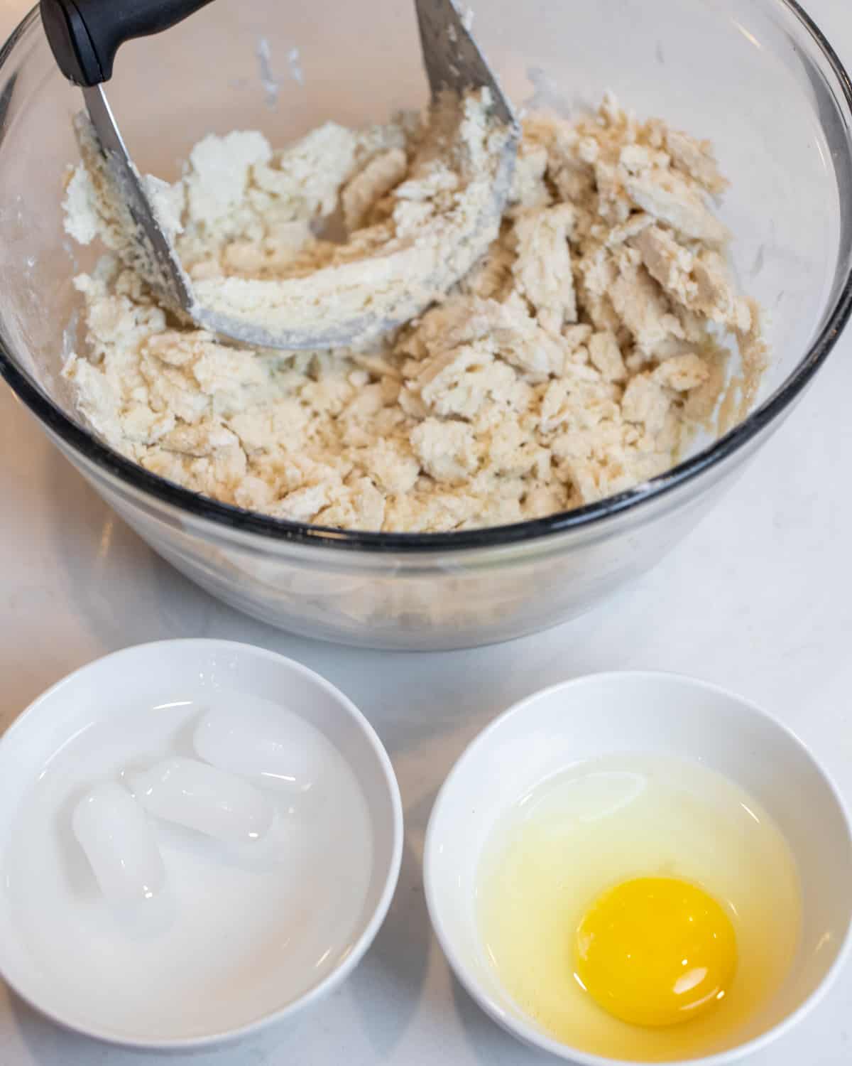 A bowl of pie dough with a small bowl with a raw egg and another bowl with water and ice cubes.